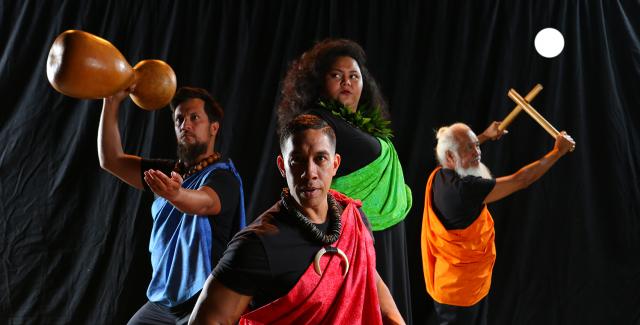In front of a black curtain, four folks in different colored satin sashes pose. Two pose with their drum or drumsticks.