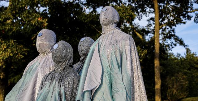 Four people wear bejeweled costumes that cover their faces. They pose plainly in front of trees.