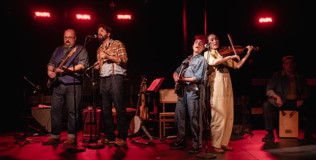 Four folks make up a band. They perform on stage, in red lights. A guitarist. A singer. A basist. A violinist.