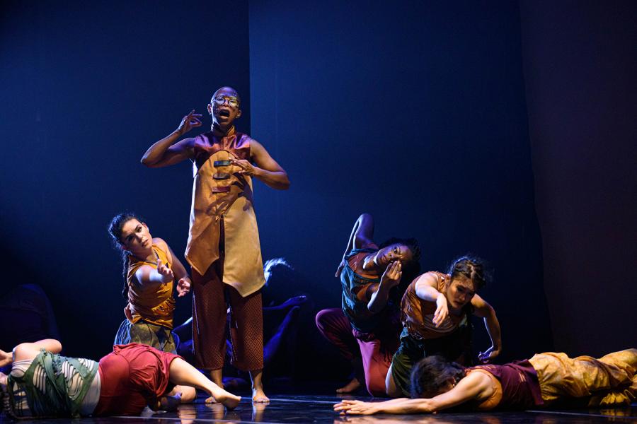 On a stage with blue light, women in red and yellow outfits roll around on the stage around a performer who stands in yellow.