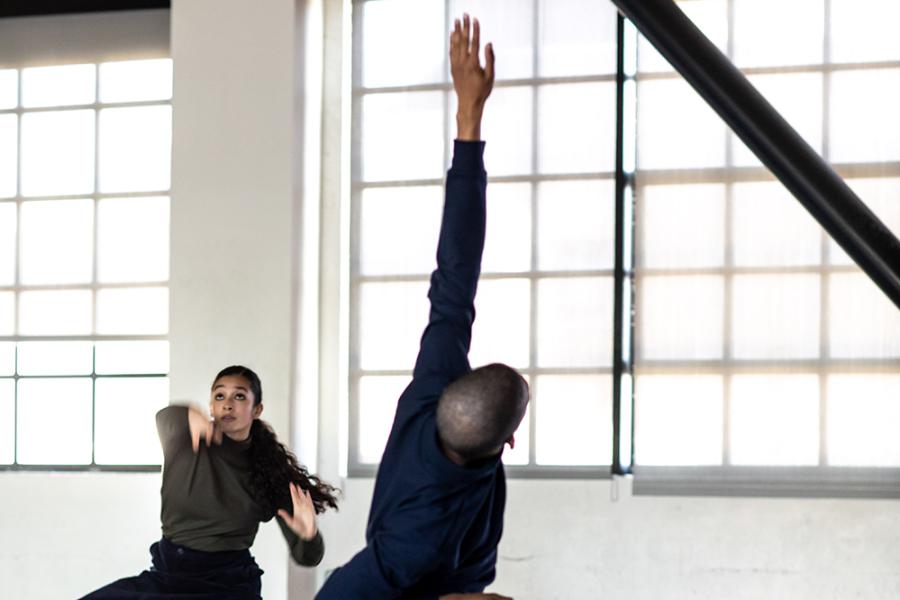 Two folks, in long black skirts, drop their legs and throw an arm up in an empty, windowed space.