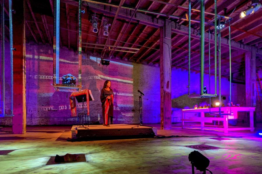 A woman, in a wrap dress, stands on a stage next to a music stand. The space has purple lights and projection of a similar fabric. It's empty otherwise.