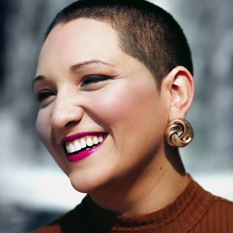 A woman with short dark hair wearing fuchsia lipstick, chunky gold earrings, and a dark red top